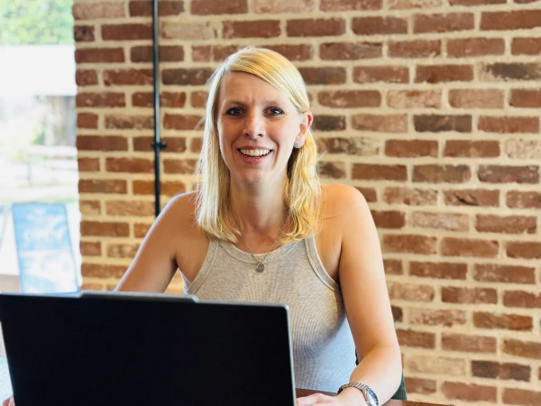 Woman with blonde hair sitting at a wooden desk with a laptop, smiling at the camera against a brick wall background