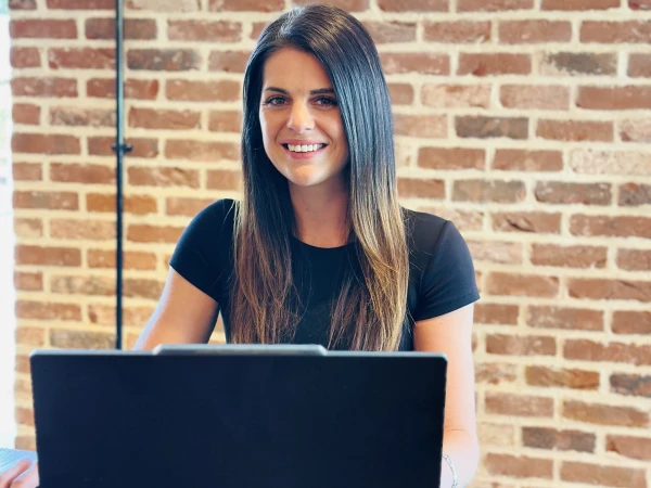 Woman with long dark hair wearing a black t-shirt, smiling at the camera whilst working on a laptop at a wooden desk, with a red brick wall behind her