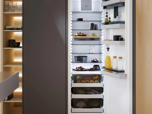 Open refrigerator with white interior displaying shelves stocked with fresh produce, bottles, jars and food items, positioned in modern kitchen with dark cabinetry and illuminated wooden storage shelving to the left