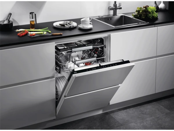 Modern kitchen with open dishwasher displaying clean dishes and utensils, black countertop with fresh vegetables, white dinnerware and cups, stainless steel sink, and minimalist décor on white cabinetry.