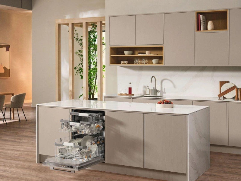 Modern kitchen with integrated dishwasher in centre island, white cabinetry with wooden accents, sink and stovetop along back wall, open shelving with decorative items, and living area visible in background