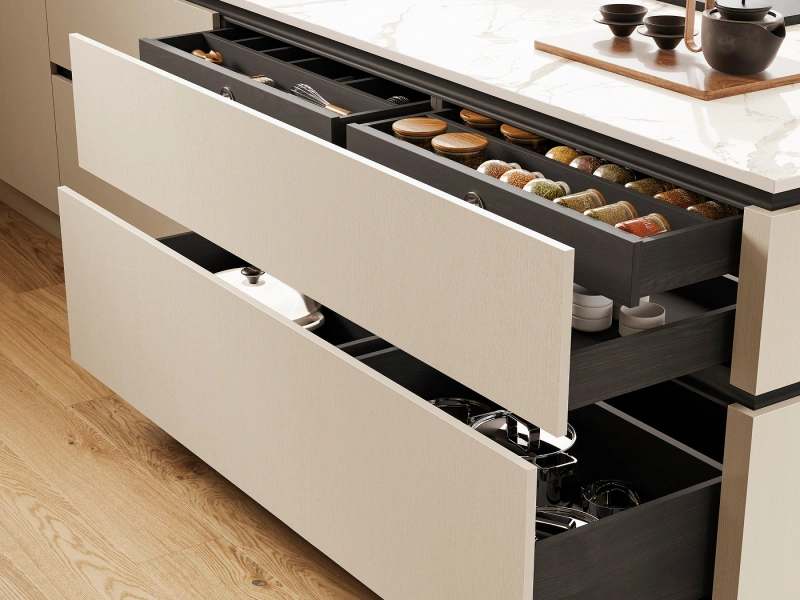 Modern kitchen with open drawers displaying organised storage including spice containers, wooden coasters, cookware and utensils, white marble countertop with dark kettle and bowls, and wooden flooring