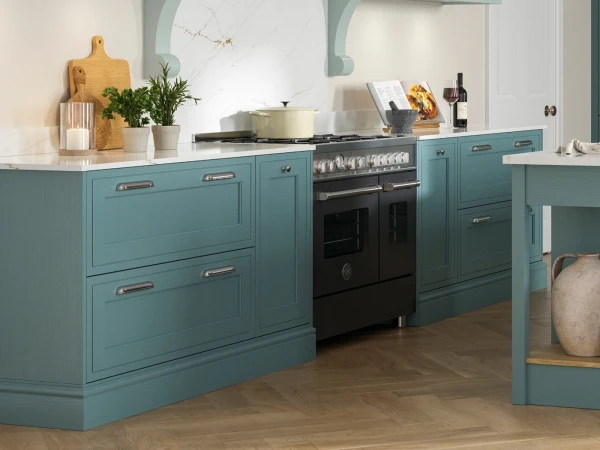 Modern kitchen with teal blue cabinetry, white marble-effect countertop, black double oven with gas hob, wooden chopping boards, potted herbs, wine bottle, cookbooks and decorative items on the counter, and herringbone wood flooring.