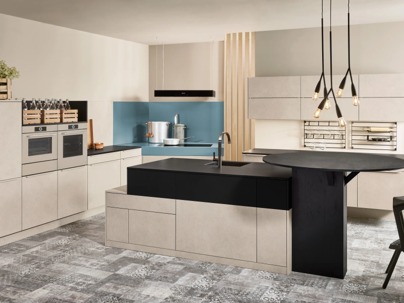 Modern kitchen interior featuring cream and black cabinetry with a central island with black countertop, blue backsplash, stainless steel appliances, pendant lighting with black cords, wooden storage crates on upper shelves, and patterned floor tiling.