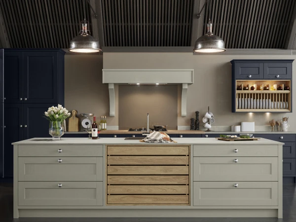 Modern kitchen with cream and dark blue cabinetry, central island featuring light green and natural wood drawers, integrated cooktop with dark surface, two pendant lights with metallic shades, white fireplace surround, open shelving displaying kitchenware and crockery, corrugated ceiling, white flowers in glass vase and various cooking utensils on counter