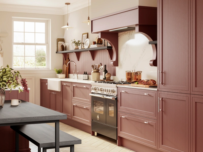 Modern kitchen with mauve-pink cabinetry, white marble splashback with curved design feature, cream walls, large window, black dining table with bench, stainless steel range cooker, pendant lights, open shelving with decorative items and potted plants.