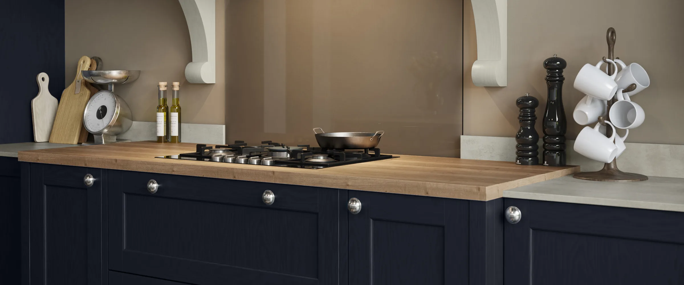 Modern kitchen with dark navy blue cabinetry, wooden countertop, black gas hob with wok, stainless steel scale, wooden chopping boards, oil bottles, black pepper mill and salt grinder, white mug tree, white paper towel holder, and beige-taupe tiled splashback with white architectural moulding details