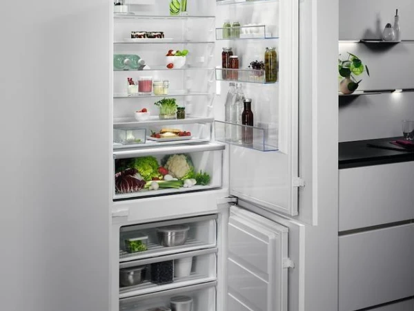 Open white built-in refrigerator with freezer compartment at the bottom, displaying fresh vegetables, bottles, jars and food items on glass shelves, positioned in a modern kitchen with grey cabinetry and dark countertop