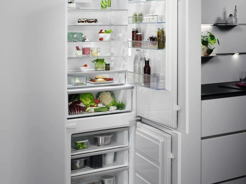 Open white built-in refrigerator with freezer compartment at the bottom, displaying fresh vegetables, bottles, jars and food items on glass shelves, positioned in a modern kitchen with grey cabinetry and dark countertop