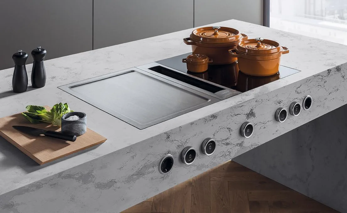 Modern kitchen island with marble countertop featuring an induction cooktop, three copper cookpots, wooden chopping board with fresh vegetables, salt and pepper mills, and control knobs along the front edge, set against grey cabinetry and bright window backdrop