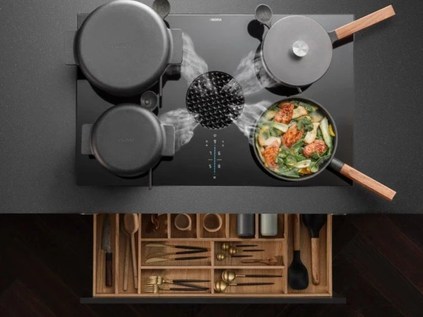 Overhead view of an induction cooktop with stainless steel pots and a pan containing colourful vegetable stir-fry, with a wooden utensil drawer below displaying organised kitchen tools and cutlery.