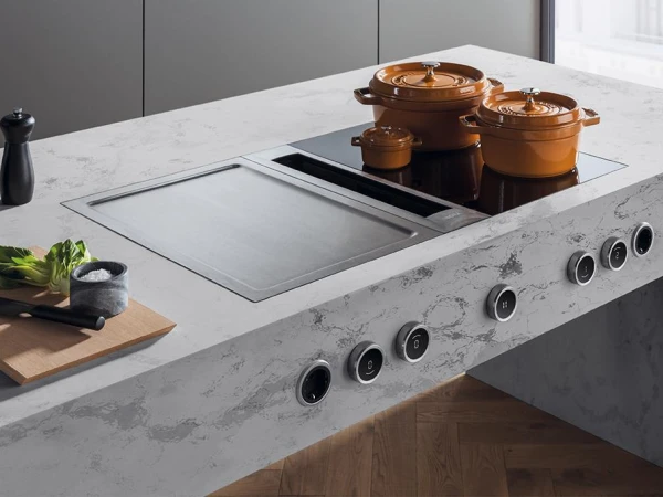 Modern kitchen island with marble countertop featuring an induction cooktop, three copper cookpots, wooden chopping board with fresh vegetables, salt and pepper mills, and control knobs along the front edge, set against grey cabinetry and bright window backdrop