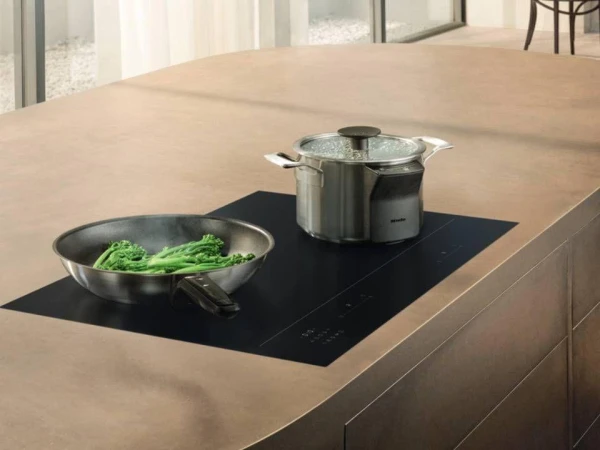 Built-in induction cooktop with a pan containing fresh greens and a stainless steel pot with lid on a light wood kitchen island, with windows visible in the background