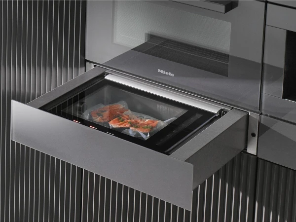 Miele steam oven drawer pulled open showing packaged salmon fillets inside the cooking chamber, with a microwave above in a modern kitchen cabinet with vertical ribbed panelling