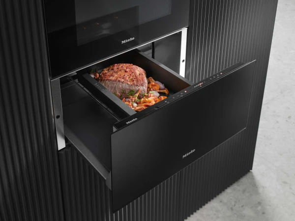 Miele built-in steam oven with open drawer displaying a cooked meat roast garnished with herbs and surrounded by roasted vegetables on a dark tray, integrated into a black kitchen cabinet unit.