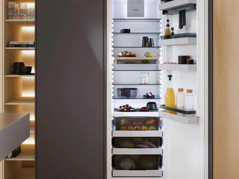 Open built-in refrigerator with white interior displaying fresh produce, bottles, jars and food items on multiple shelves, positioned within a modern kitchen with dark cabinetry and illuminated wooden shelving unit on the left side