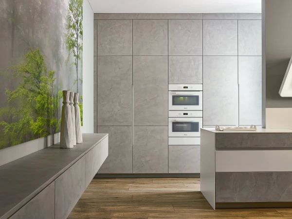 Modern minimalist kitchen with grey stone cabinetry, built-in white appliances including two ovens, wooden flooring, white cloth napkins in a vase on the counter, and a living green plant wall feature on the left side