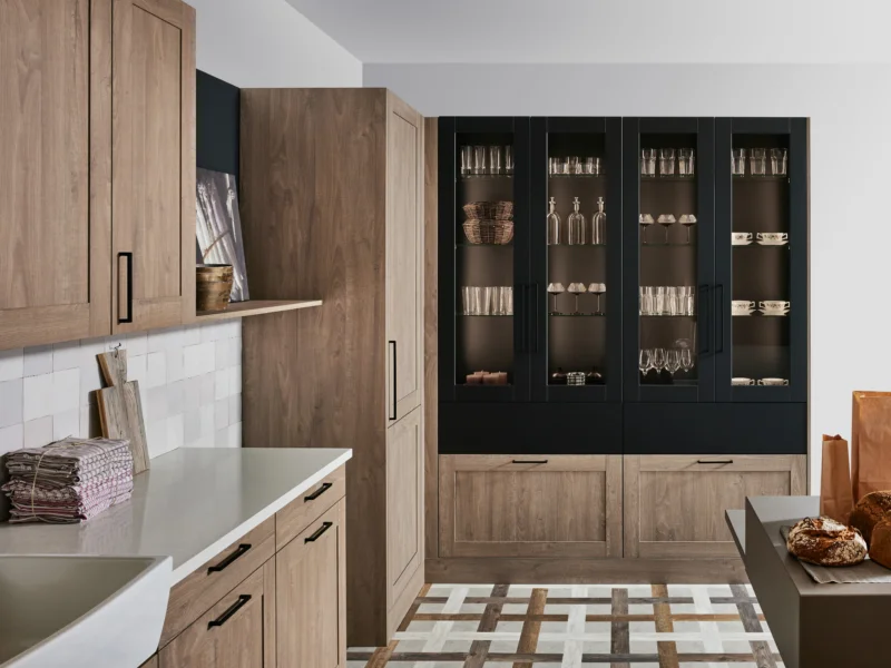 Modern kitchen showroom display with warm wood cabinetry, white countertops, black glass-fronted display units, and geometric floor tiling.