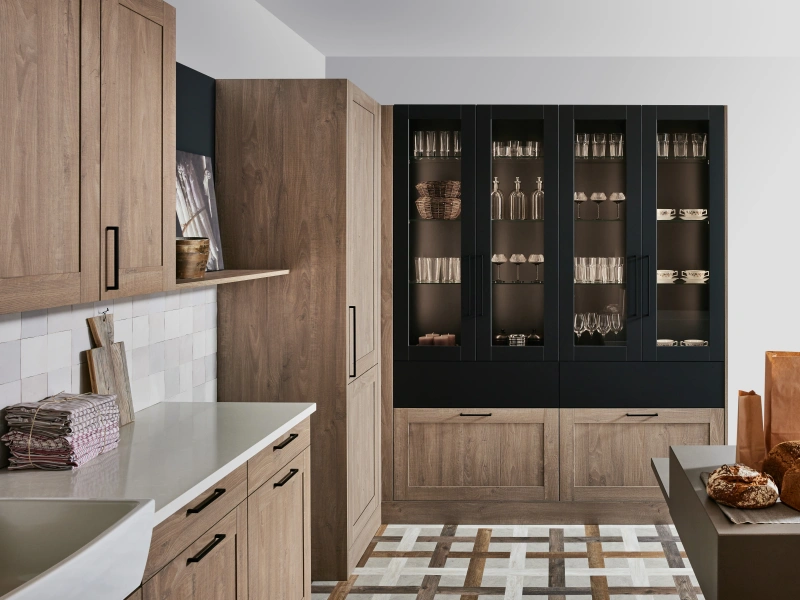 Modern kitchen showroom display with warm wood cabinetry, white countertops, black glass-fronted display units, and geometric floor tiling.