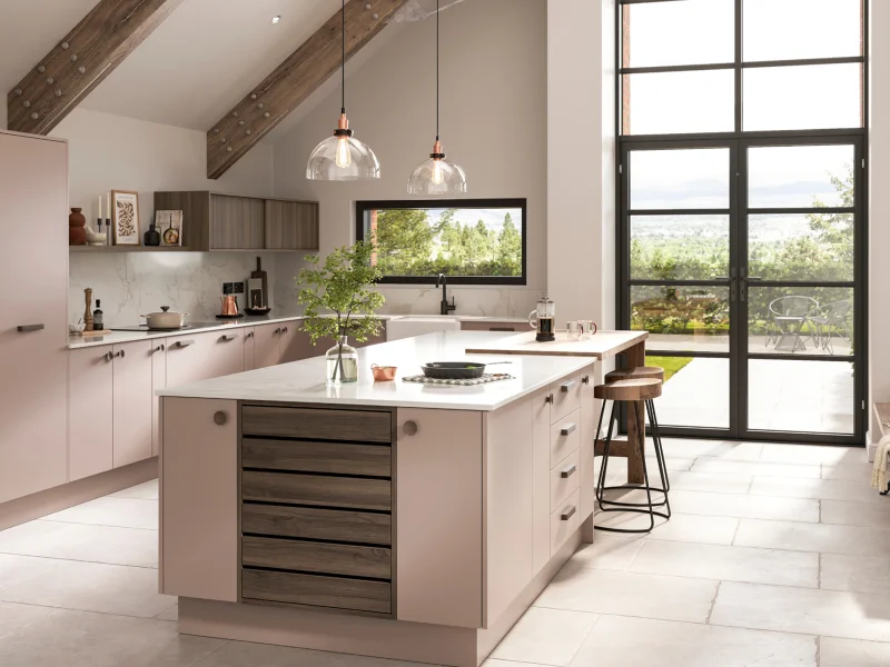 Modern open-plan kitchen and dining area with pink cabinetry, central island with white worktop, exposed wooden beams, pendant lights, black framed windows and doors overlooking countryside, dining table with place settings on the right, and seating area by the window.