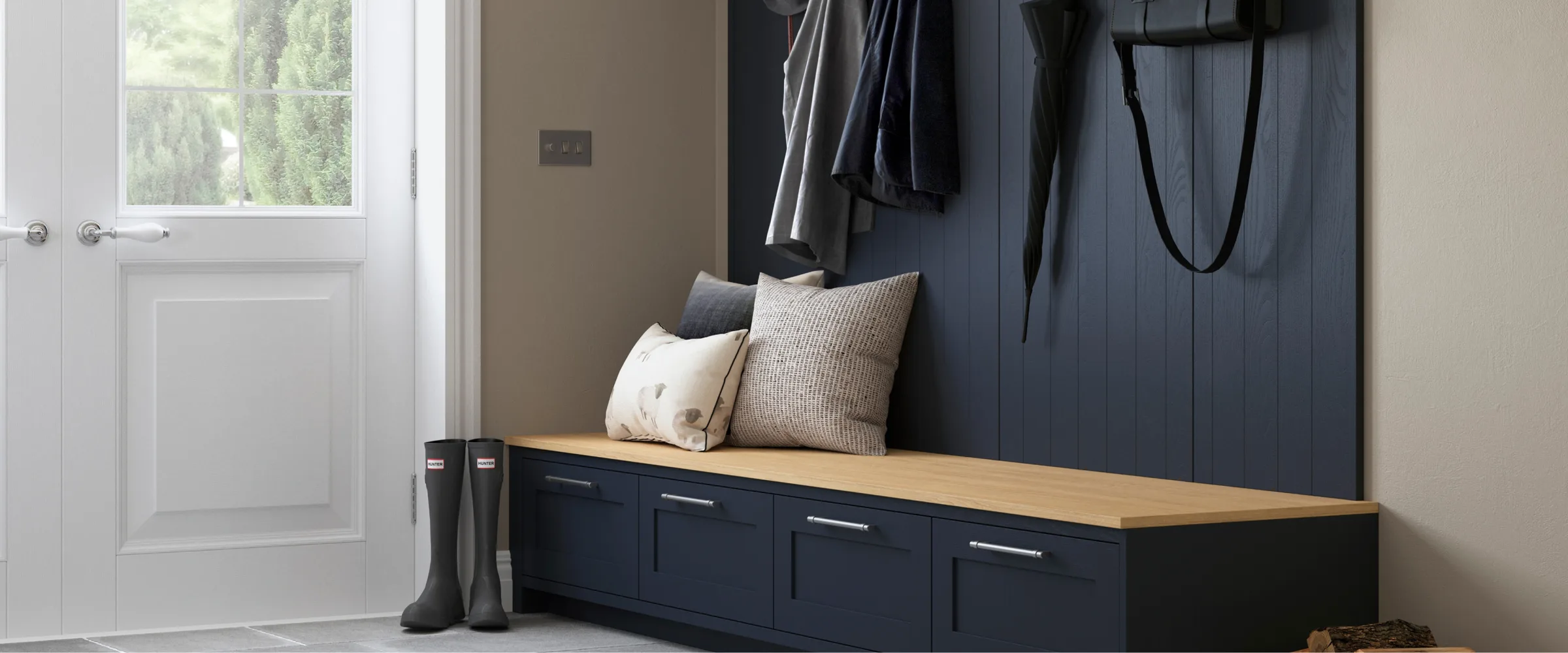 Modern entryway with a dark blue storage bench featuring a natural wood top, decorative cushions, and metal handles, positioned against a dark blue panelled wall with clothing hanging above, white door with window to the left, black boots on the floor, and neutral beige walls.
