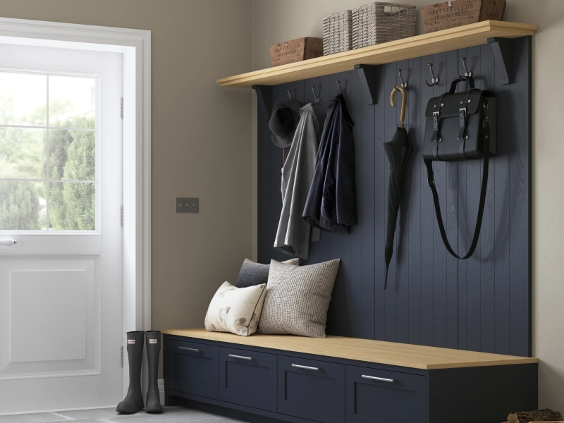 Modern entryway with navy blue shaker-style wall panelling and matching storage bench with natural wood top, featuring hooks for hanging coats and bags, upper shelving with baskets and storage boxes, cream cushions on the bench seat, white door with glass panels leading outside, and black boots on the floor.