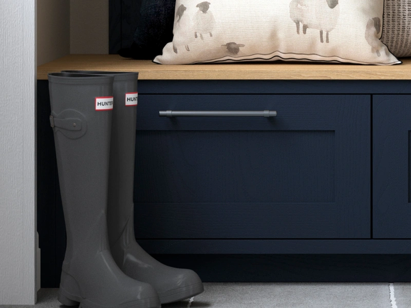 Dark grey Hunter wellington boots on a floor beside a navy blue storage bench with a wooden top, displaying a cream cushion with sheep pattern and folded textiles in the background.