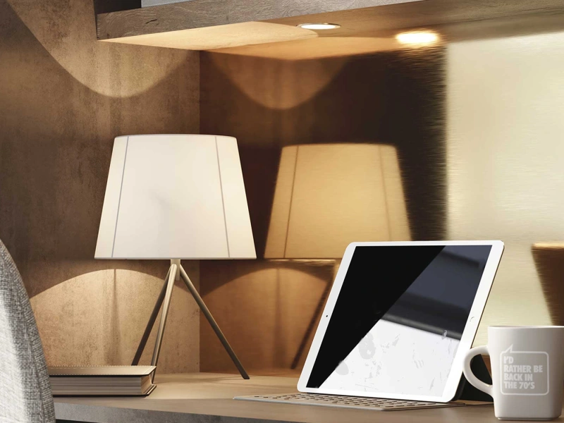 Modern workspace shelf displaying a white table lamp, beige fabric lampshade, tablet device, wireless keyboard, and mug beneath wooden beams with integrated lighting