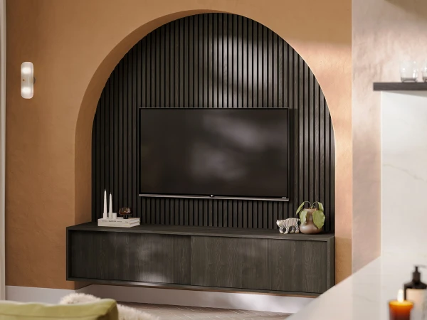 Modern living room with a large arched alcove featuring a flat-screen television mounted on vertical black slat panelling, dark wood media console below with decorative items including books, a zebra figurine and potted plant, warm beige walls with cylindrical wall sconce lighting, and partial views of furniture and home accessories in the foreground