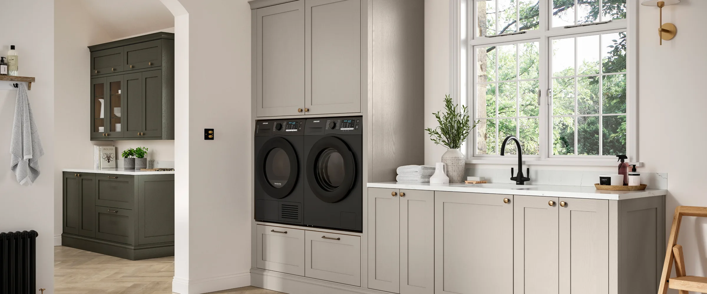 Modern laundry room with pale grey cabinetry, black integrated washer and dryer, white countertop with black faucet, large multi-pane window overlooking greenery, potted plant, folded towels, and dark green cabinetry visible in adjoining room