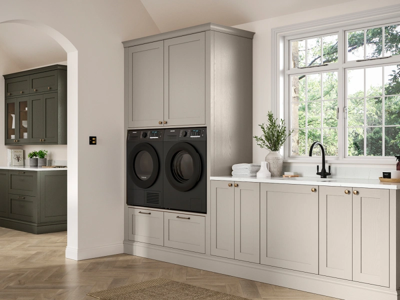 Modern laundry room with cream-coloured cabinetry, two black front-load washing machines stacked vertically, white countertop with sink and black tap, large multi-pane window overlooking greenery, potted plant and rolled white towels, dark green cabinetry visible in adjacent kitchen area, herringbone wood flooring and neutral beige walls.