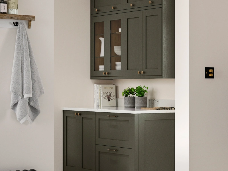 Bathroom vanity with olive green shaker-style cabinetry featuring brass knob handles, white countertop with potted plants and decorative items, glass-fronted upper cabinets displaying white dishware, grey towel hanging from wooden shelf, and black light switch on cream walls.