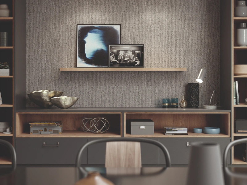 Modern home office with textured grey wallpaper, wooden floating shelf displaying two framed artworks, open shelving units with decorative objects and books, desk with chair in foreground, and integrated storage cabinets below