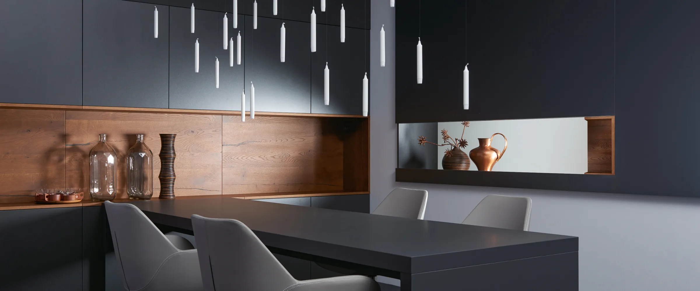 Modern kitchen interior with dark cabinetry and wooden accents, featuring a black dining table with grey chairs, hanging tubular light fixtures above, glass bottles on a wooden shelf, copper vessels displayed in a wall niche, and warm ambient lighting throughout the contemporary space.