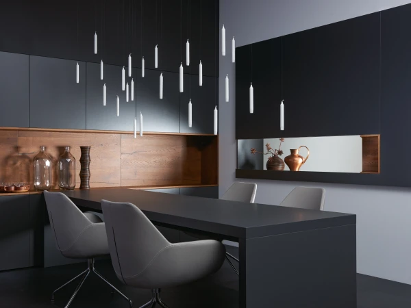 Modern kitchen and dining area with black and natural wood cabinetry, featuring a dark island with grey upholstered chairs, decorative hanging white pendant lights, glass bottles and copper vessels displayed on wooden shelves, and minimalist monochromatic design throughout.