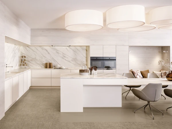 Modern minimalist kitchen and dining space with white cabinetry, marble backsplash, integrated appliances, central island with waterfall countertop, cylindrical pendant lights, grey dining chairs, and decorative accessories including amber glass vases and dried flowers.
