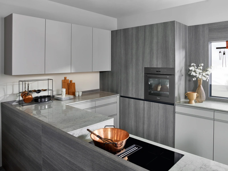 Modern kitchen featuring grey wood-effect cabinetry and white upper cabinets, with a central island topped in light grey marble and equipped with an induction hob, built-in black oven, stainless steel countertop, and copper cookware, decorated with fresh white flowers in a vase and wooden kitchen utensils.