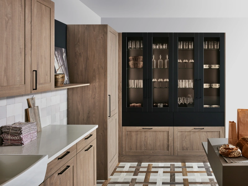 Modern kitchen interior with natural wood cabinetry, white countertops and subway tile backsplash on the left, and a tall black display cabinet with glass doors on the right containing glassware and decorative items, geometric patterned floor in neutral tones