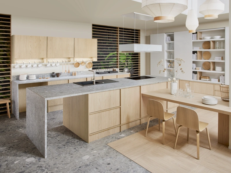 Modern open-plan kitchen and dining space with light wood cabinetry, grey concrete countertops, white shelving units, pendant lighting, and a wooden dining table with two chairs, featuring floor-to-ceiling windows with black louvred blinds overlooking greenery