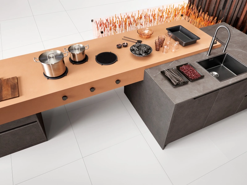Overhead view of a modern modular kitchen setup with a copper-coloured cooking unit featuring stovetop burners, pots and pans, and a charcoal grey sink and prep unit with faucet, displayed on white tiled flooring with decorative burnt orange wall art in the background.