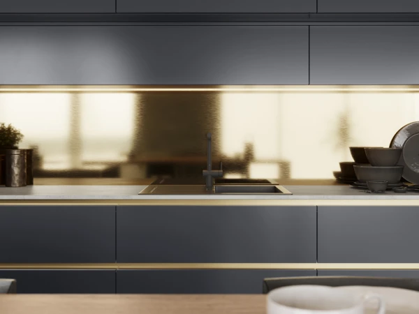 Modern kitchen interior with dark grey cabinetry, brass accents, and a cream-coloured mirrored backsplash. A sink with dark tap is centred on the counter, flanked by a potted plant on the left and stacked bowls on the right. A white mug sits in the foreground.