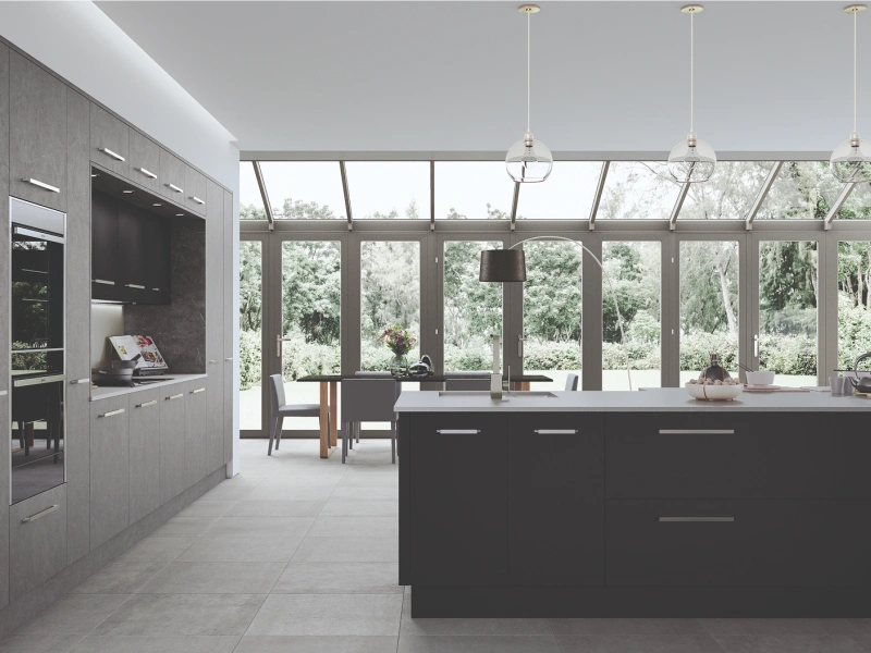 Modern kitchen with grey and black cabinetry, large island with white countertop, pendant lights, and extensive glazed conservatory-style roof overlooking green woodland views, featuring built-in appliances and dining area with chairs