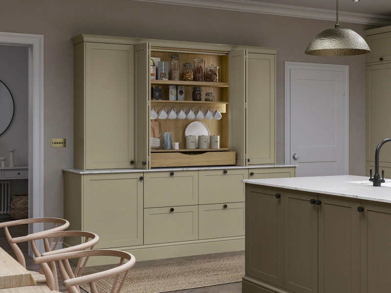 Contemporary kitchen with sage green and cream cabinetry, featuring open shelving displaying glassware and kitchen items, white marble countertops, black hardware, a silver dome pendant light, wooden dining chairs in the foreground, and white panelled doors on the walls.
