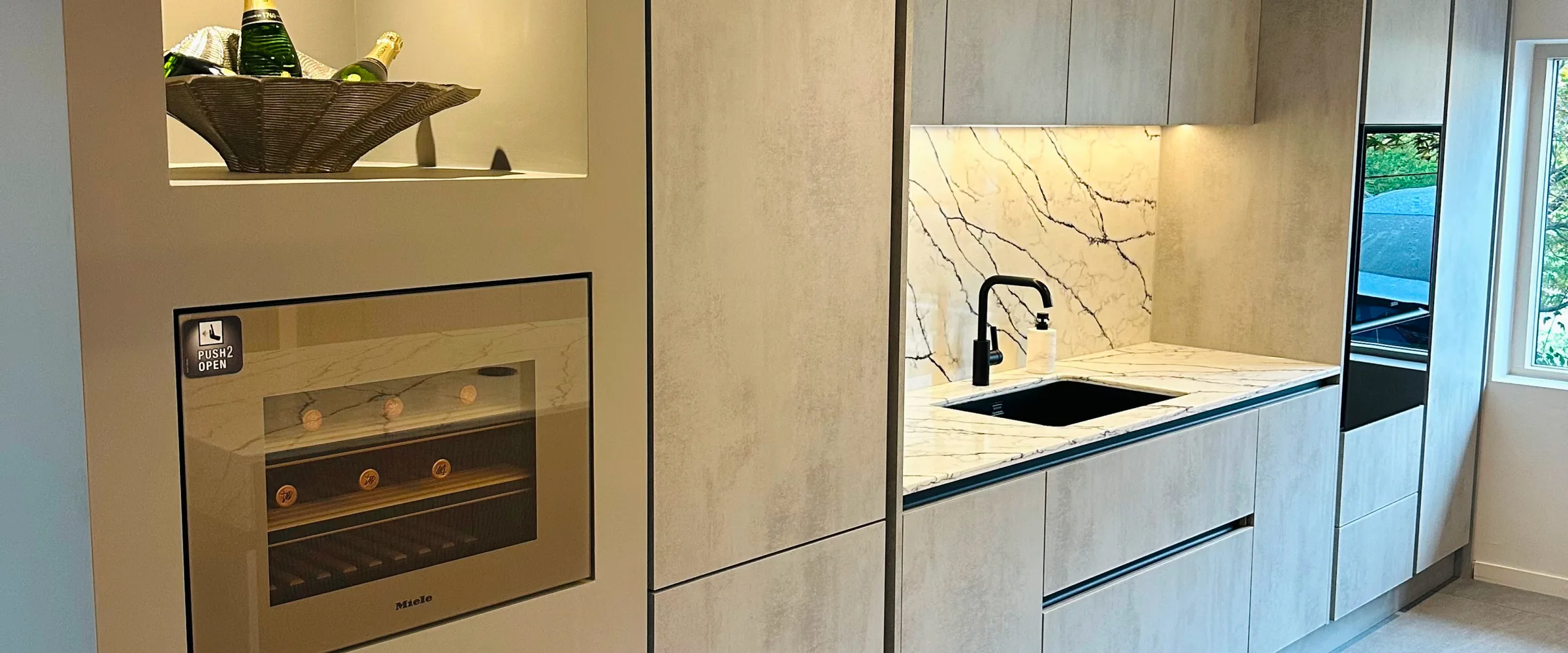 Modern kitchen interior with cream and grey cabinetry, featuring a built-in Miele oven on the left with a wine cooler above, a central island with marble countertop and black sink with faucet, and a backlit mirror panel, with water views visible through glass doors on the right