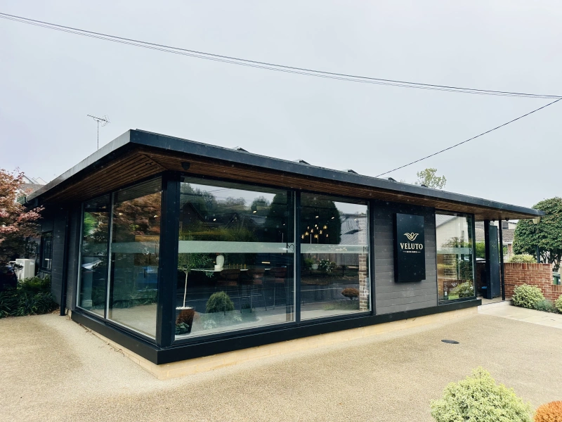 Modern black pavilion building with large glass windows and wooden interior ceiling, featuring Veluto branding on the storefront, situated on a gravel forecourt with surrounding gardens and overcast sky
