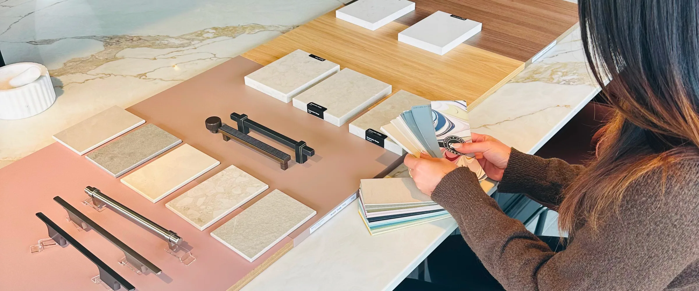 Person viewing kitchen and bathroom design samples displayed on a countertop, including cabinet hardware, tile samples in neutral tones, and wood flooring options.