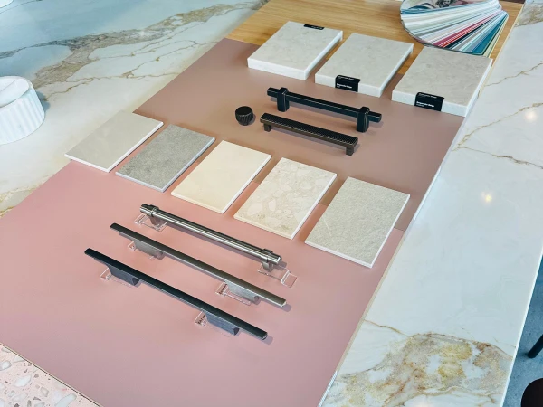 Interior design showroom display featuring tile and material samples arranged on pink and tan boards, with black cabinet hardware handles and a colour palette fan swatch on a wooden table, set against marble-effect flooring.