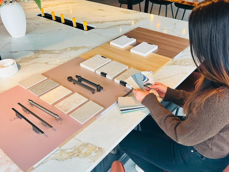 Person reviewing material and hardware samples displayed on a marble table in a modern showroom, with colour swatches, cabinet handles, and decorative accessories arranged for selection.