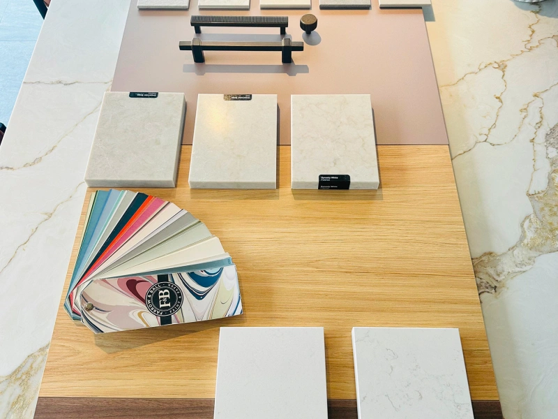 Overhead view of a kitchen and bathroom design showroom display featuring tile and flooring samples arranged on a wooden platform, including marble effect tiles, paint colour swatches, wood finish samples, and display hardware tools against marble-effect wall backgrounds.